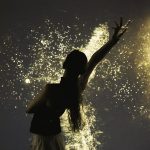 A silhouetted figure reaches upward (dancing) while surrounded by cascading golden projected pollen particles, creating a dramatic contrast against a dark background.