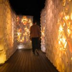 A person walking through a narrow illuminated corridor lined with honeycomb-patterned panels glowing in warm shades of amber and gold.